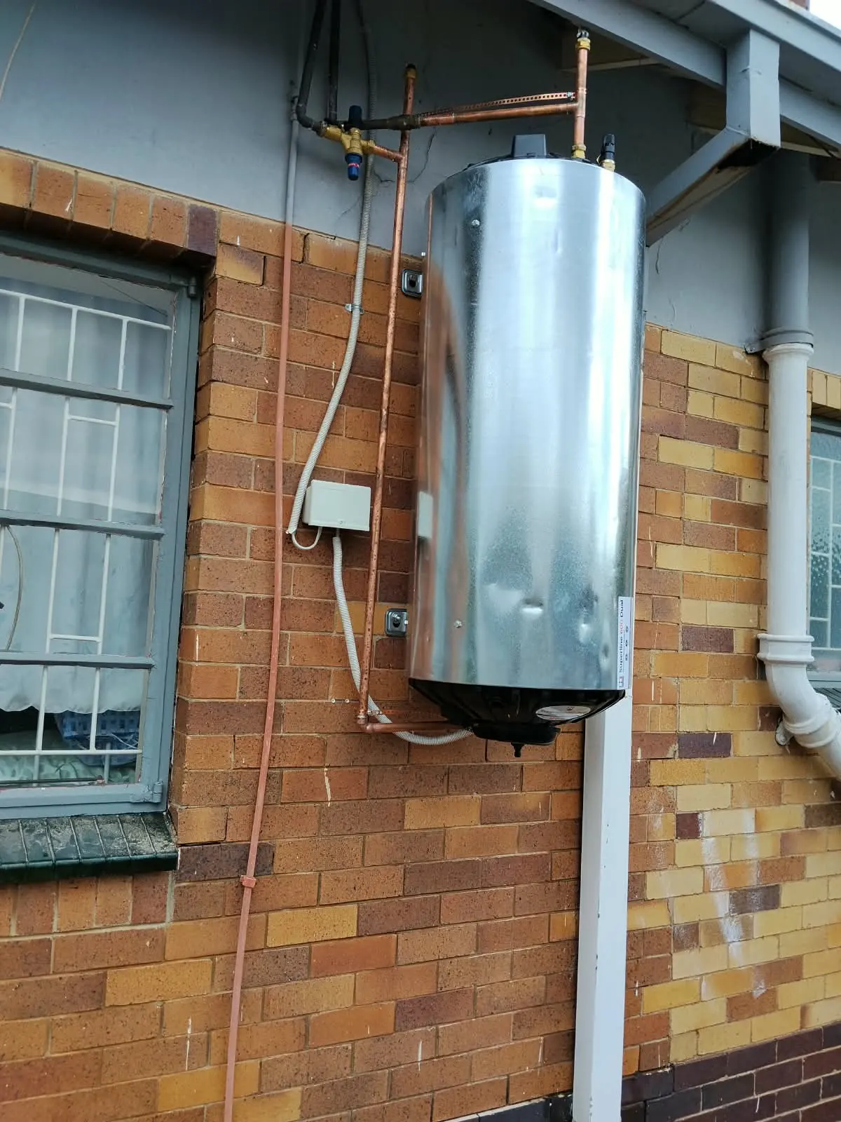 Vertical geyser mounted on an outside brick wall with copper pipes and electrical conduit