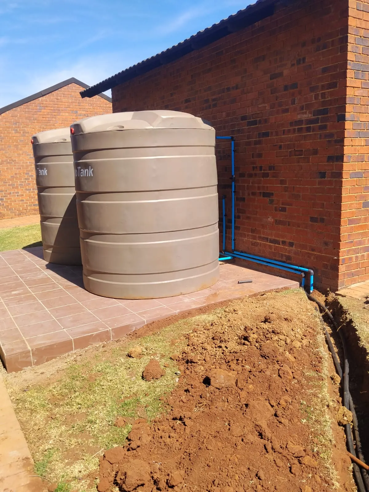 Two JoJo water tanks installed on a paved base beside a brick house, connected with blue water pipes